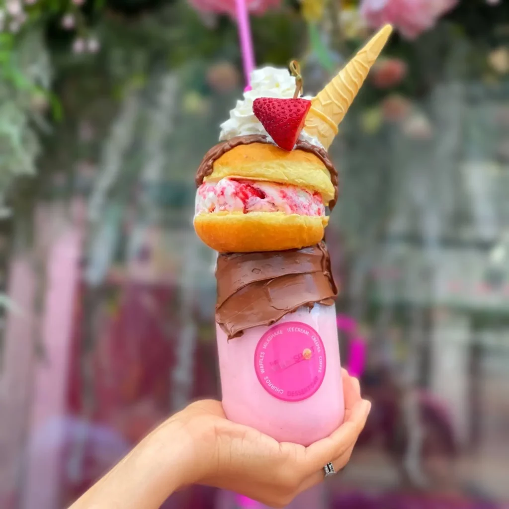 A hand holds a mason jar with a strawberry shortcake milkshake and a Nutella rim. A doughnut filled with strawberry ice cream. Topped with whipped cream, strawberries, and a waffle cone.